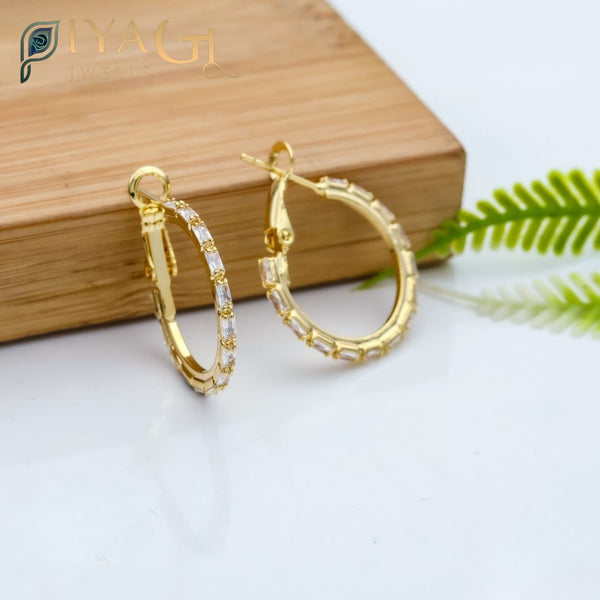 Gold-colored hoop earrings with baguette cut stones resting on a wooden block. - PiyaGuJwells