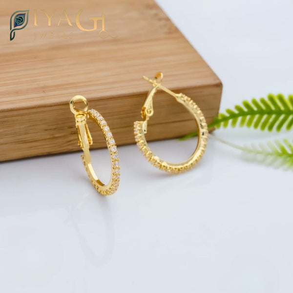A pair of gold-colored square hoop earrings with pave-set clear stones, displayed on a wooden surface next to greenery. - PiyaGuJwells