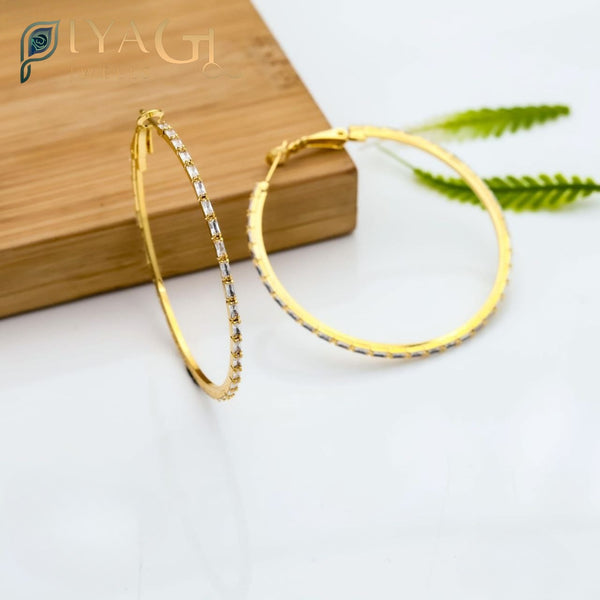 A pair of large gold-colored hoop earrings with clear stones set along the rim, displayed on a wooden surface. - PiyaGuJwells