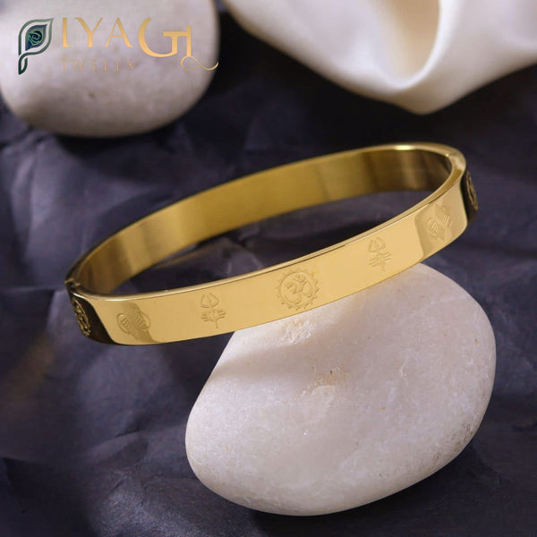 Golden stainless steel bangle with engraved symbols resting on a white stone. - PiyaGuJwells