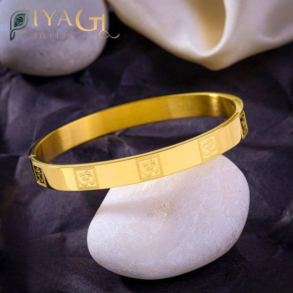Gold-colored stainless steel bangle bracelet with embossed Om symbols resting on a smooth white stone. - PiyaGuJwells