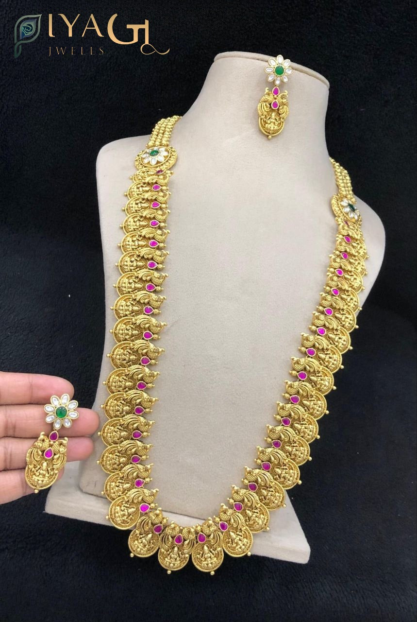 Woman wearing an antique gold finish long necklace with red and green stones, matching earrings, traditional Indian imitation jewelry set - PiyaGuJwells